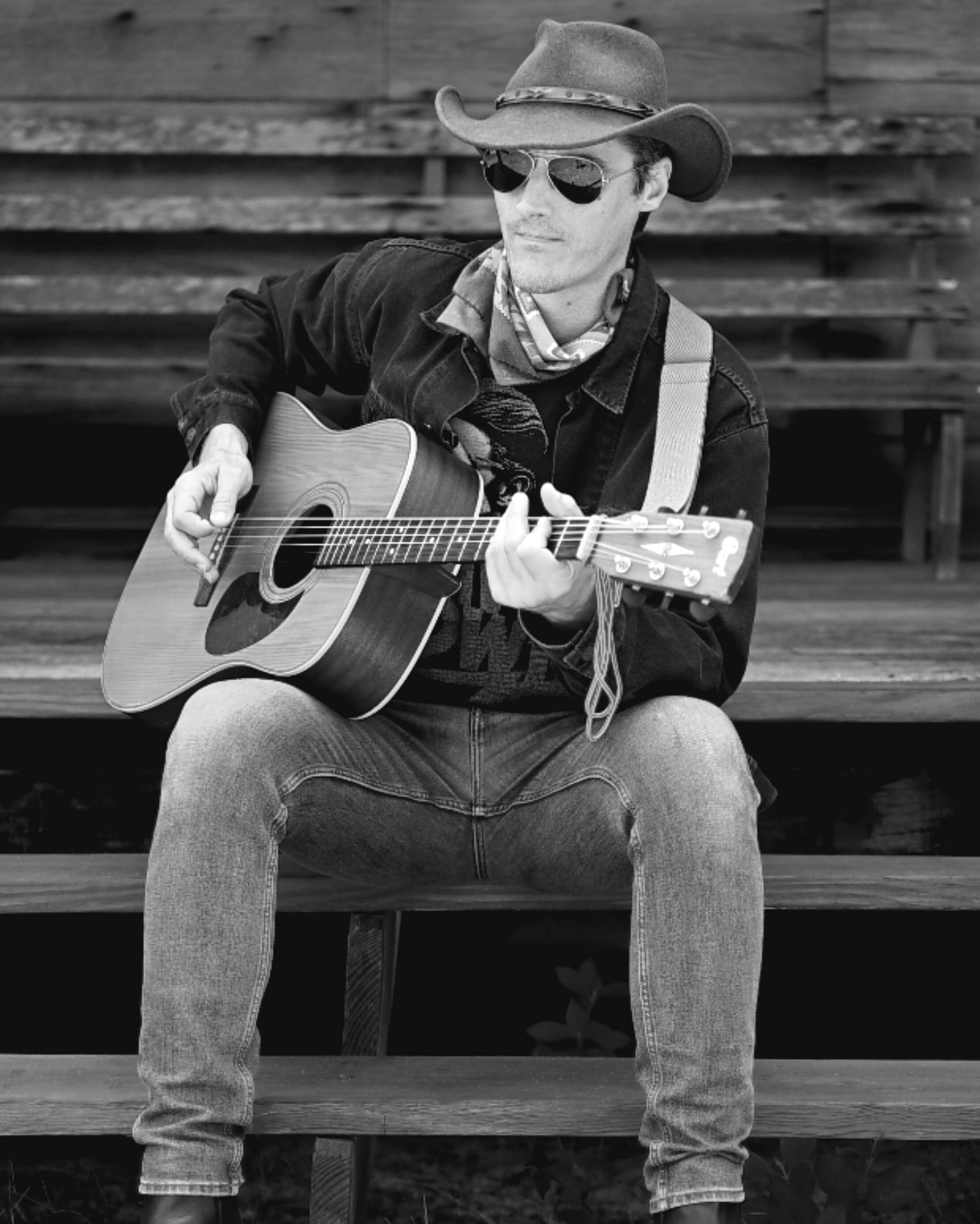 Justin on the porch A man dressed as a cowboy sits on a rustic porch playing a guitar. Photo by Dylan Jon Wade Cox.